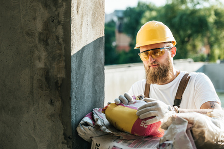builder in protective goggles and hardhat opening bag of cement at construction siteの写真素材