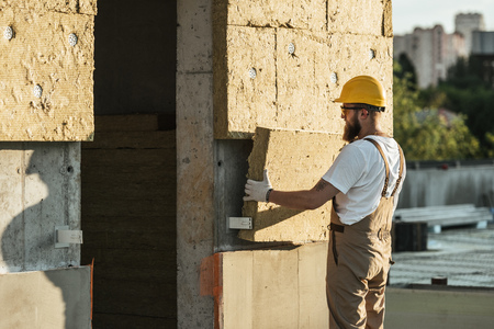 side view of builder in protective helmet and goggles doing insulation of wall at construction site