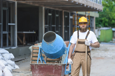 young builder in hardhat and protective goggles standing with shovel near concrete mixer at...