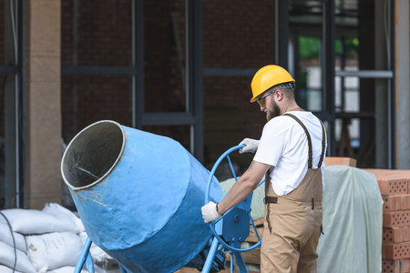 construction worker in hardhat and protective goggles working with concrete mixer on construction...
