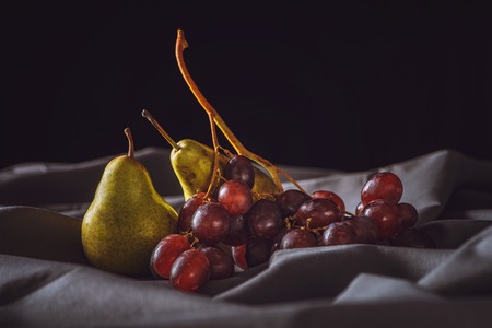 close-up shot of ripe pear and red grapes on grey drapery on blackの写真素材