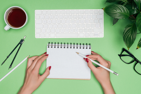 cropped image of female architect writing in empty textbook at table with computer keyboard, divider and coffee cupの写真素材