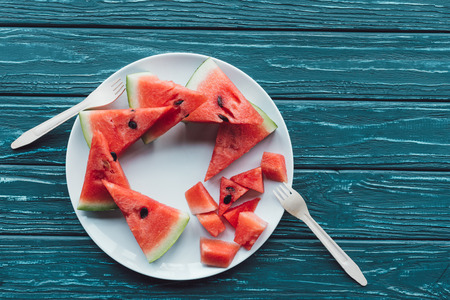 flat lay with arranged watermelon pieces on plate and plastic forks on blue wooden surfaceの写真素材