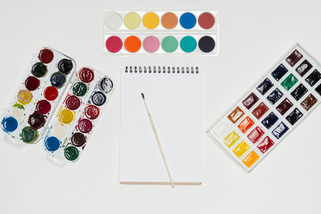 top view of empty textbook and paintbrush surrounded by colorful paints on white tableの写真素材