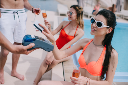 cropped shot of smiling girl paying with credit card while drinking champagne with friends at poolの写真素材