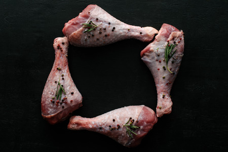flat lay with uncooked chicken legs with pepper corns and rosemary on black wooden tableの写真素材