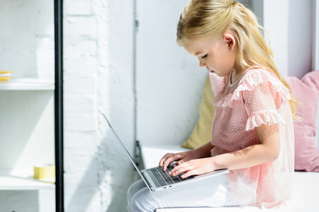 side view of cute little child using laptop while sitting on windowsill