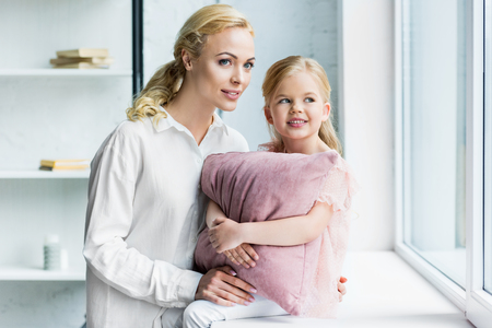 mother and adorable smiling daughter with pillow looking at window at homeの写真素材