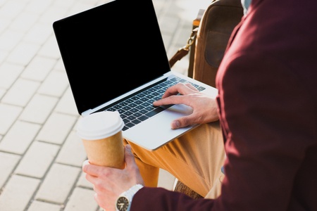 cropped shot of man holding paper cup and using laptop with blank screen on streetの写真素材