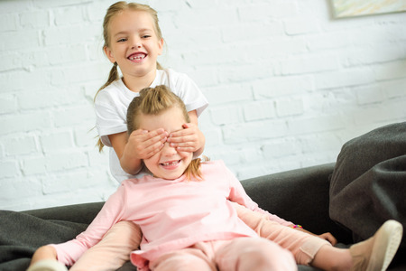 adorable little kid covering eyes to sister on sofa at homeの写真素材