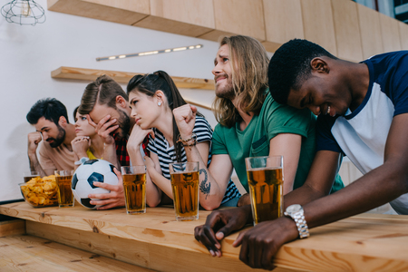 upset multicultural group of friends sitting at bar counter and watching football matchの写真素材