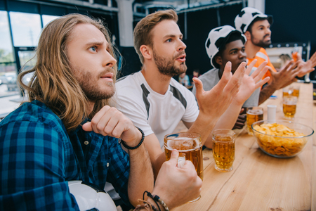 upset multicultural group of male football fans in soccer ball hats drinking beer and watching football match at barの写真素材