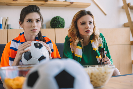 focused female football fans in different t-shirt and scarfs watching soccer match at homeの写真素材