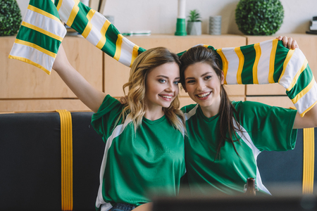 smiling female football fans in green t-shirts and scarf celebrating during watch of soccer match at homeの写真素材