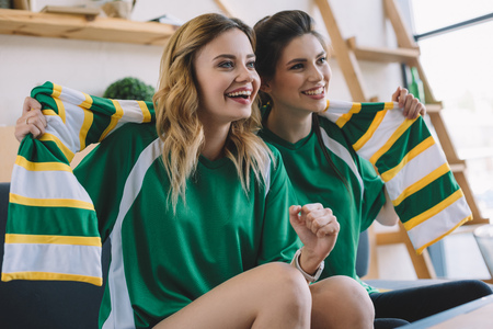happy female football fans in green t-shirts and scarf gesturing by hands during watch of soccer match at homeの写真素材