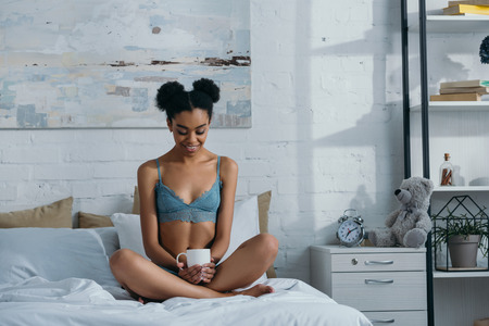 smiling african american girl in underwear with cup of coffee sitting on bed in bedroom in morningの写真素材