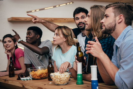 two men pointing by hands to friends sitting at bar with fan horns, popcorn, chips and beer bottles during watch of football matchの写真素材