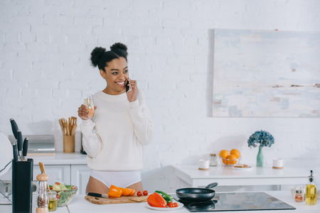 beautiful young woman with glass of champagne preparing breakfast and talking by phone at homeの写真素材