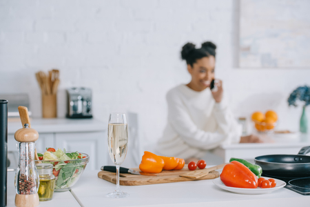 blurred smiling young woman talking by phone with vegetables and glass of champagne on foreground at homeの写真素材