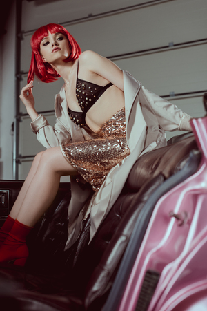 low angle view of stylish girl in red wig looking away while sitting on vintage carの写真素材