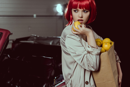 stylish girl in trench coat holding paper bag with lemons and looking awayの写真素材