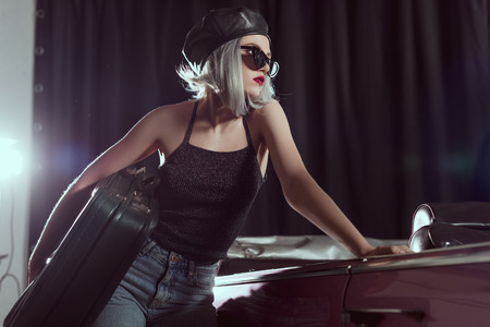 beautiful young woman in sunglasses and beret holding vintage suitcase and looking away while leaning at classic carの写真素材