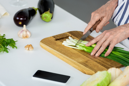 cropped image of grey hair woman cutting green onion on wooden board in kitchenの写真素材