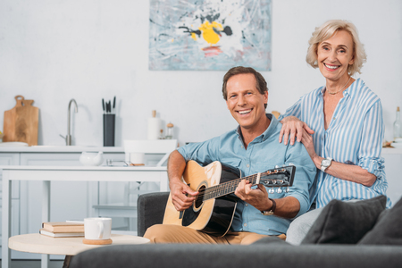 happy senior couple smiling at camera while man playing guitar at homeの写真素材