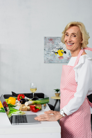 side view of smiling attractive senior woman using laptop for cooking in kitchenの写真素材