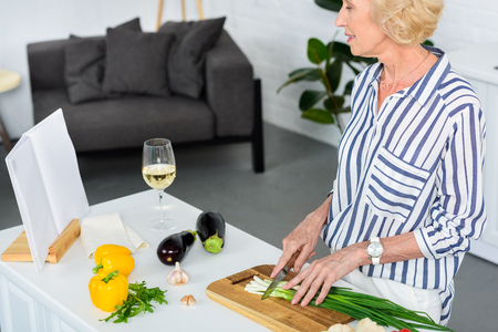 attractive grey hair woman cutting green onion in kitchen and looking at recipe bookの写真素材