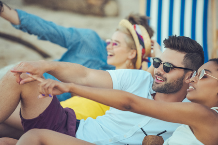 selective focus of multiracial smiling friends pointing away while resting on beachの写真素材