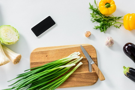 green onion on wooden board, smartphone with blank screen on white tabletop in kitchenの写真素材