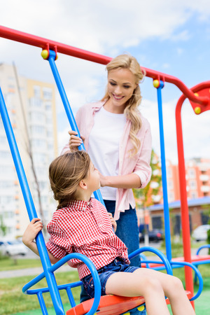 selective focus of smiling mother riding daughter on swing at playgroundの写真素材