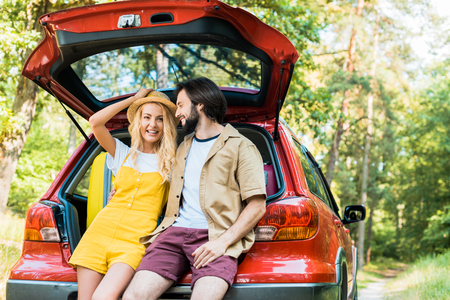 smiling couple sitting on car trunk in forestの写真素材