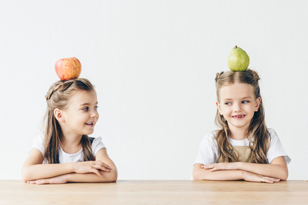 happy little schoolgirls with apple and pear on heads sitting at table isolated on whiteの写真素材