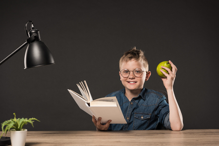 smiling schoolboy holding pear and reading book at table with plant and lamp on grey backgroundの写真素材