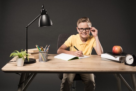 little schoolboy in eyeglasses doing homework at table with books, plant, lamp, colour pencils, apple, clock and textbook on grey backgroundの写真素材