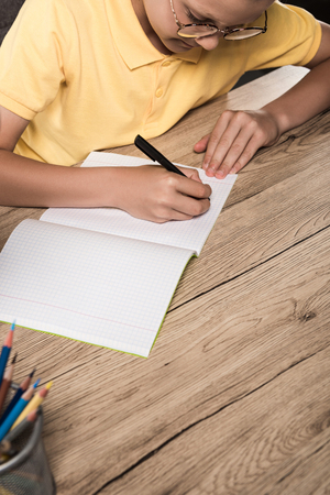 partial view of schoolboy in eyeglasses writing in empty textbook at table with colour pencilsの写真素材