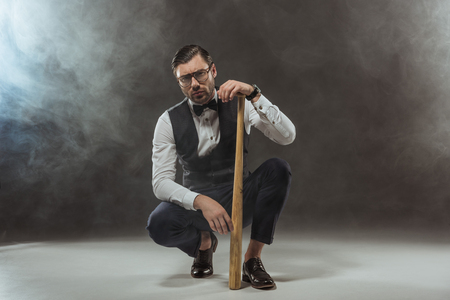 serious stylish man in eyeglasses crouching with baseball bat and looking at camera on blackの写真素材
