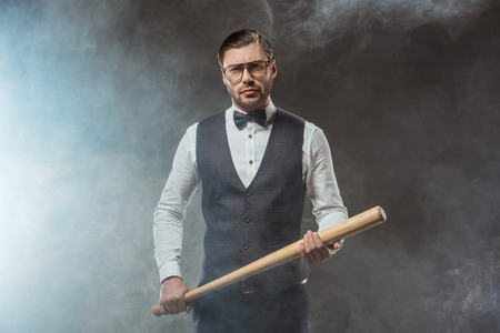handsome man in bow tie and eyeglasses holding baseball bat and looking at camera in smokeの写真素材