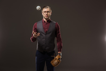 serious businessman with baseball glove throwing ball and looking at camera isolated on blackの写真素材