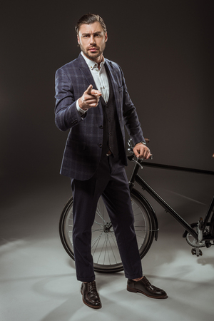 serious young man in suit pointing at camera while standing near bicycle on blackの写真素材