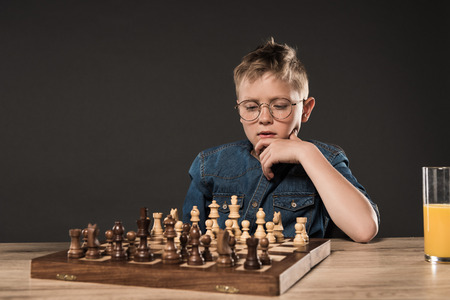 pensive little boy sitting at table with chess board and glass of juice on grey backgroundの写真素材