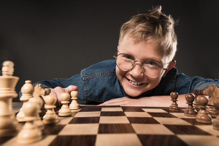 smiling little boy in eyeglasses looking at camera while sitting at table with chess board isolated on grey backgroundの写真素材