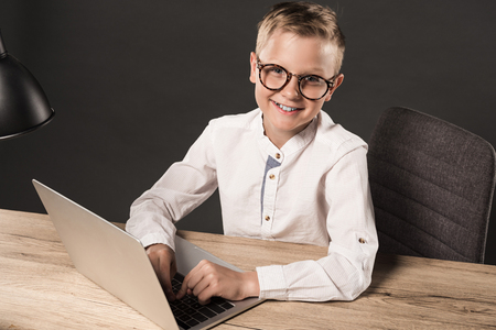 happy little boy in eyeglasses looking at camera while sitting at table with laptop and lamp on grey backgroundの写真素材