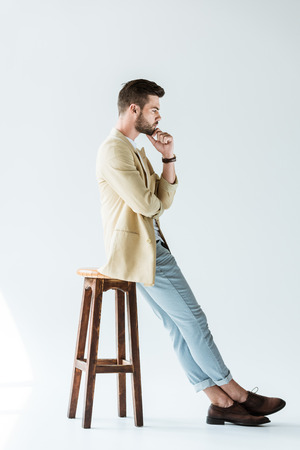 Stylish young man thinking while sitting on stool on white backgroundの写真素材
