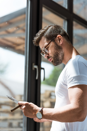 Fashionable confident man holding cigar and reading report by windowの写真素材