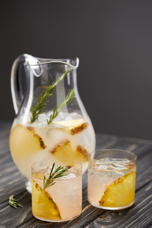 jug and two glasses of lemonade with pineapple pieces, ice cubes and rosemary on grey wooden tabletopの写真素材