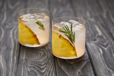 two glasses of lemonade with pineapple pieces, ice cubes and rosemary on grey wooden tabletopの写真素材