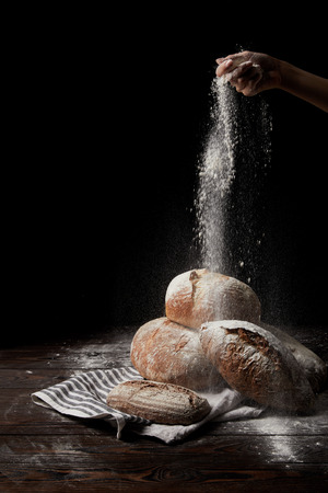 partial view of female baker sieving flour over various types of bread on sackcloth isolated on black backgroundの写真素材